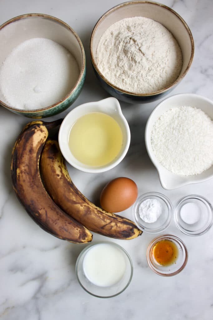 Ingredienten voor een kokos bananencake: rijpe bananen, bakjes met bloem, suiker, kokos, een los ei, glazen schaaltjes met  bakingsoda, zout, vanille extract en karnemelk