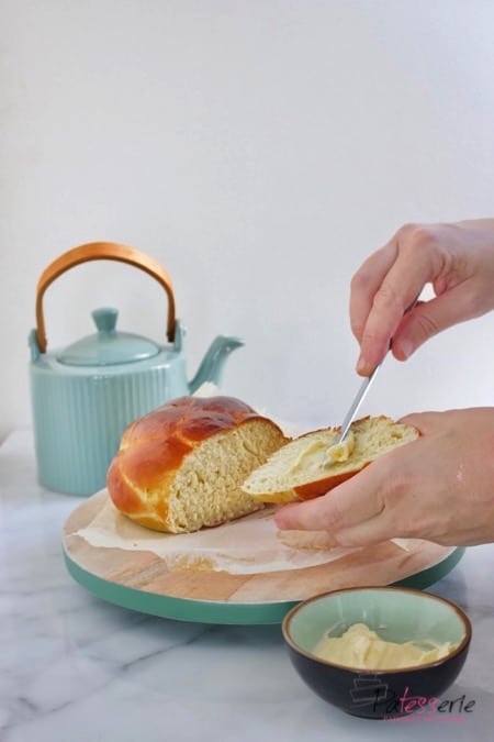 Een luchtig gevlochten aardappelbrood, wat besmeerd wordt met boter, op een snijplank met een theepot ernaast.