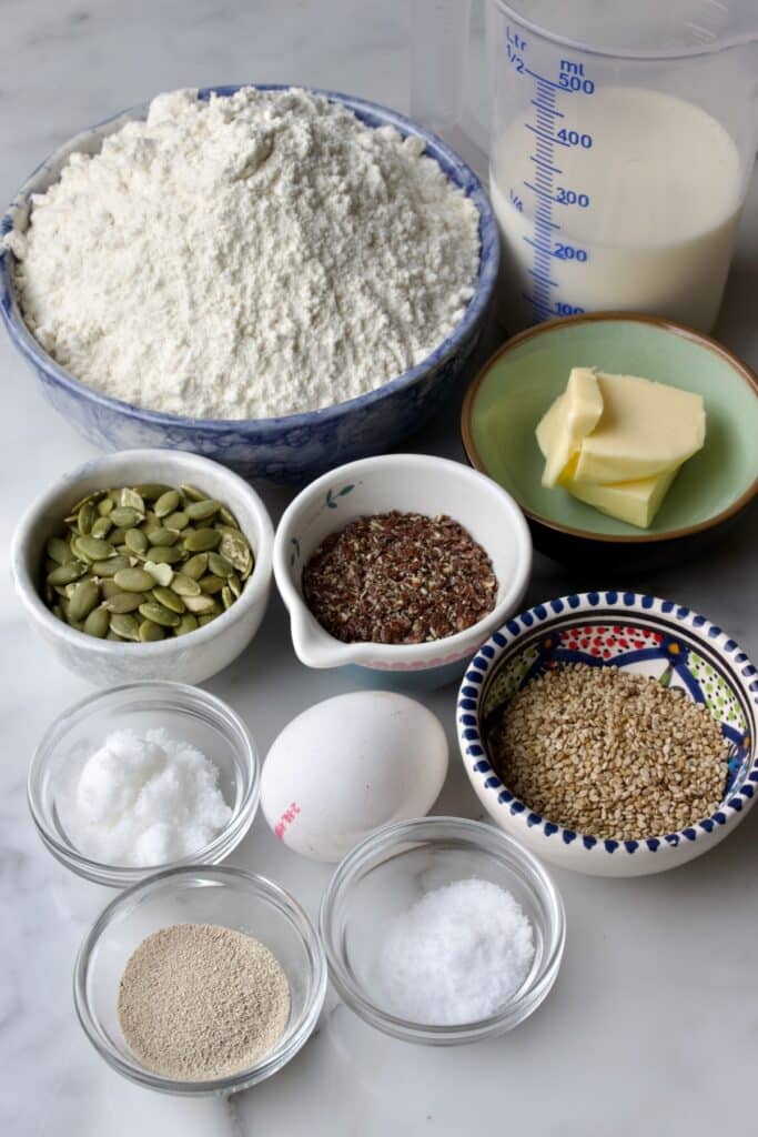 Ingredienten in schaaltjes voor het breekbrood met zaden en pitten: tarwebloem, melk, boter, suiker, zout, ei, gist, sesamzaad, lijnzaad en pompoenpitten